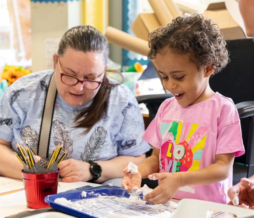 An adult and a child sit at a table. The child plays with a tray of white, foamy material and looks excited. The adult, wearing glasses, watches and guides. Pencils in a red holder and papers are also on the table.