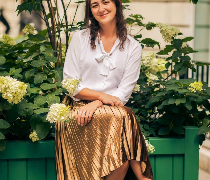 A woman with long brown hair, wearing a white blouse and gold pleated skirt, sits on the edge of a green planter box with flowering plants, smiling at the camera.