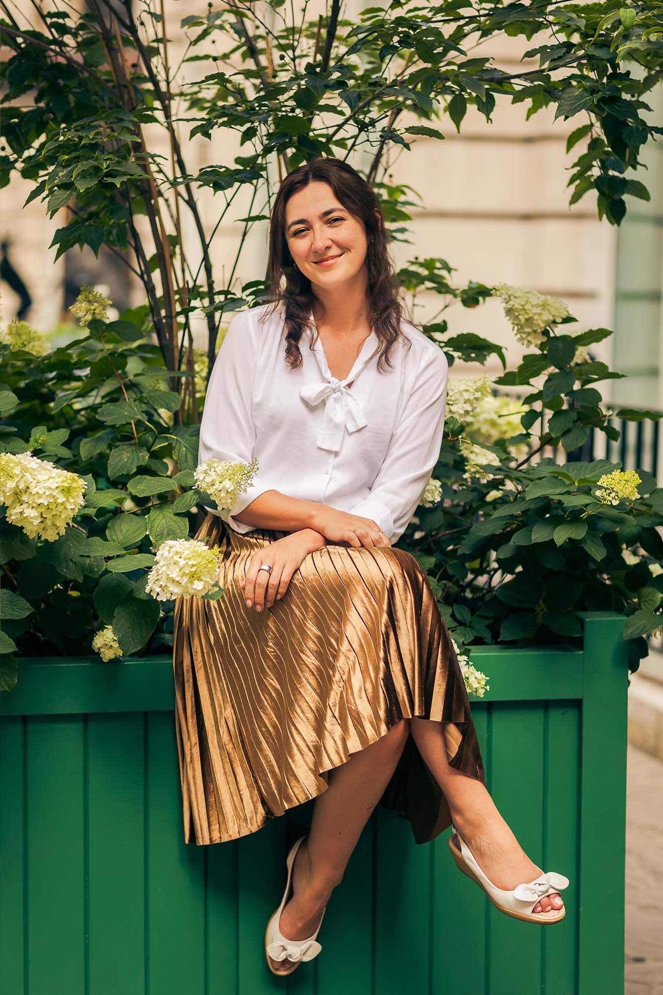 A woman with long brown hair, wearing a white blouse and gold pleated skirt, sits on the edge of a green planter box with flowering plants, smiling at the camera.