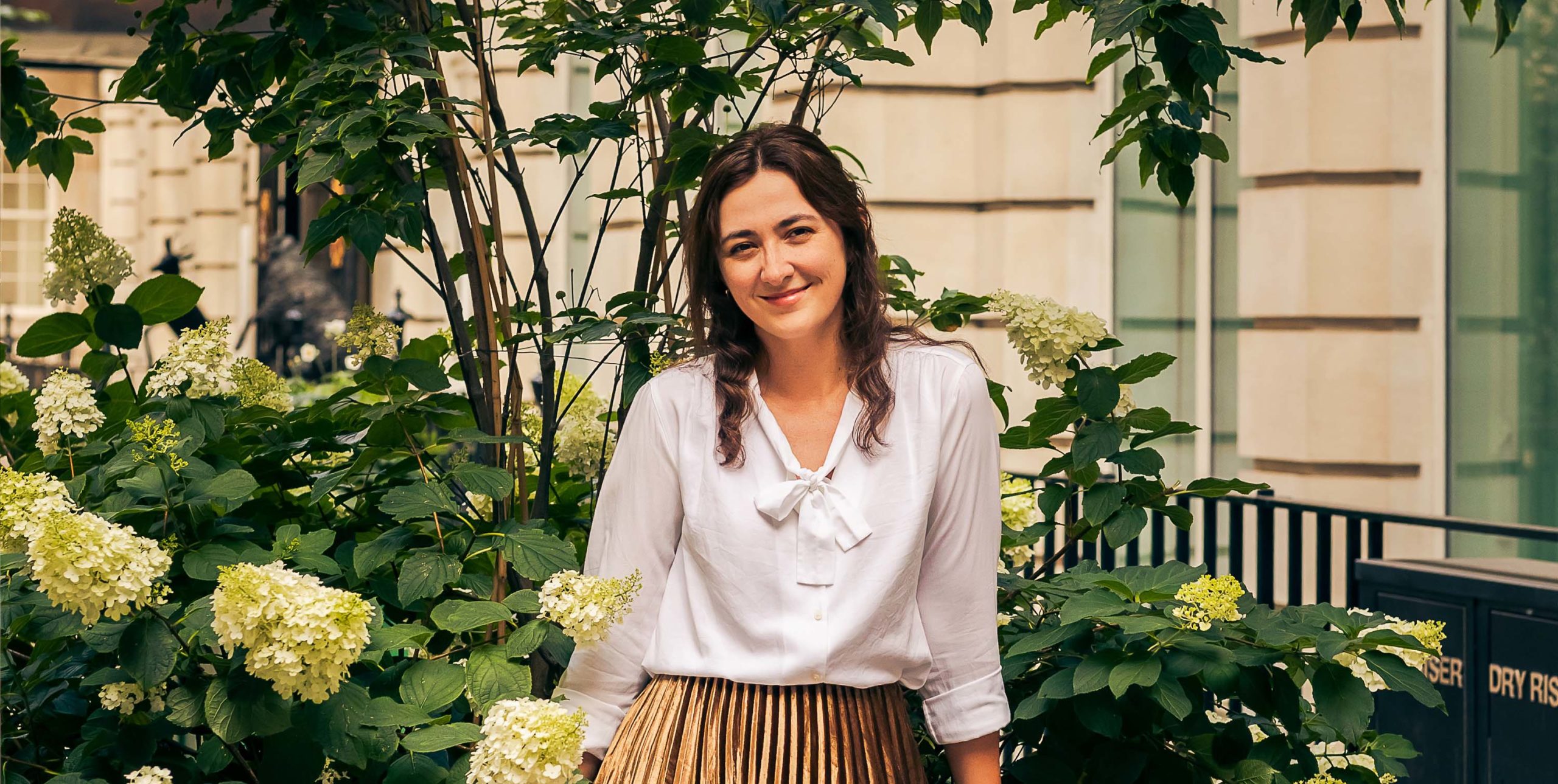 A woman with wavy brown hair stands in front of flowering hydrangea bushes, wearing a white blouse and a gold pleated skirt, with a building and railing in the background.