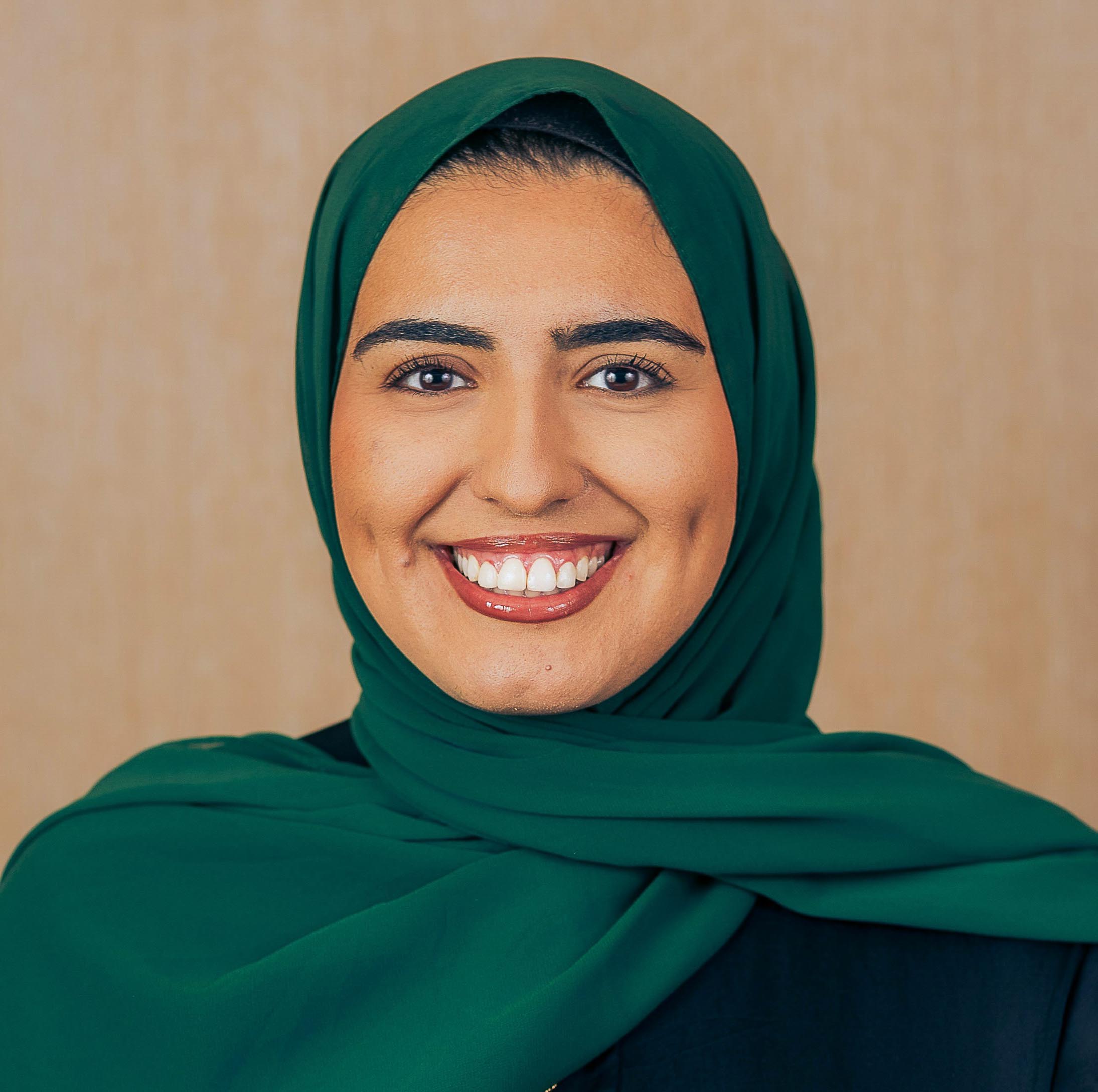 A woman wearing a dark green scarf, black top and round gold necklace, against a natural woodgrain background.