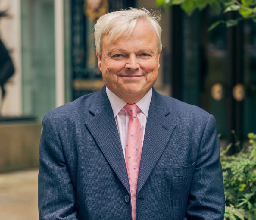A man wearing a dark blue suit, white shirt, and pink tie, stands outdoors with his hands clasped, smiling at the camera. Green plants and a building entrance are visible in the background.