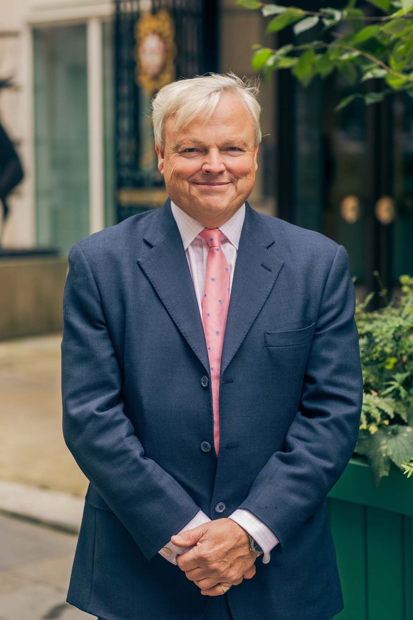 A man wearing a dark blue suit, white shirt, and pink tie, stands outdoors with his hands clasped, smiling at the camera. Green plants and a building entrance are visible in the background.