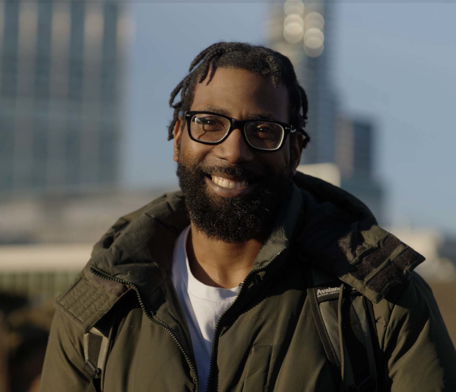 A man with glasses, a beard, smiles outdoors. He is wearing a green jacket and a rucksack. Buildings are blurred in the background.