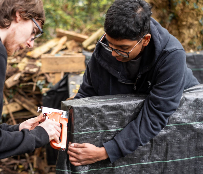 Two people work together outside, one holding a staple gun to secure black fabric onto a wooden frame whilst the other holds the fabric in place. There are trees and wooden debris in the background.