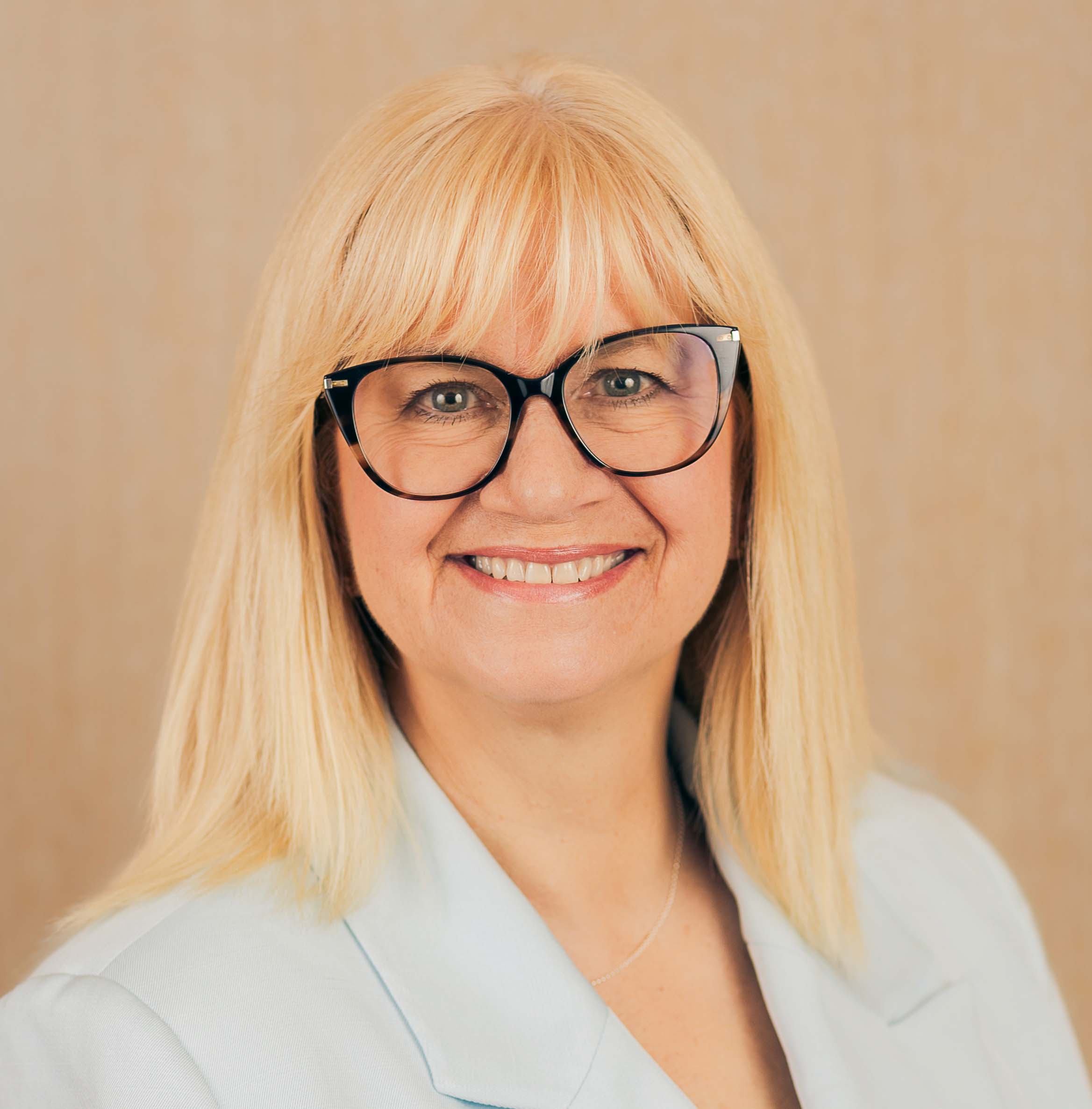 A woman with straight blonde hair and black glasses smiles at the camera. She is wearing a light blue blazer and standing in front of a neutral wood grain background.