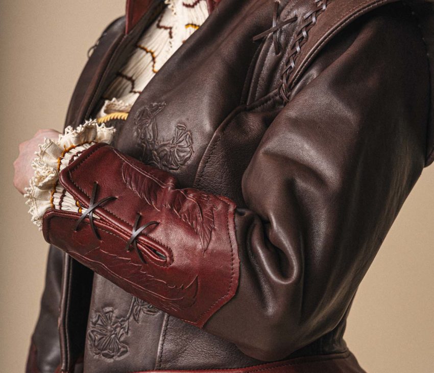 A person wears a brown leather jacket with red accents, decorative stitching, and embossed details over a cream ruffle-collared blouse. Only the lower face, torso, and part of the left arm are visible.
