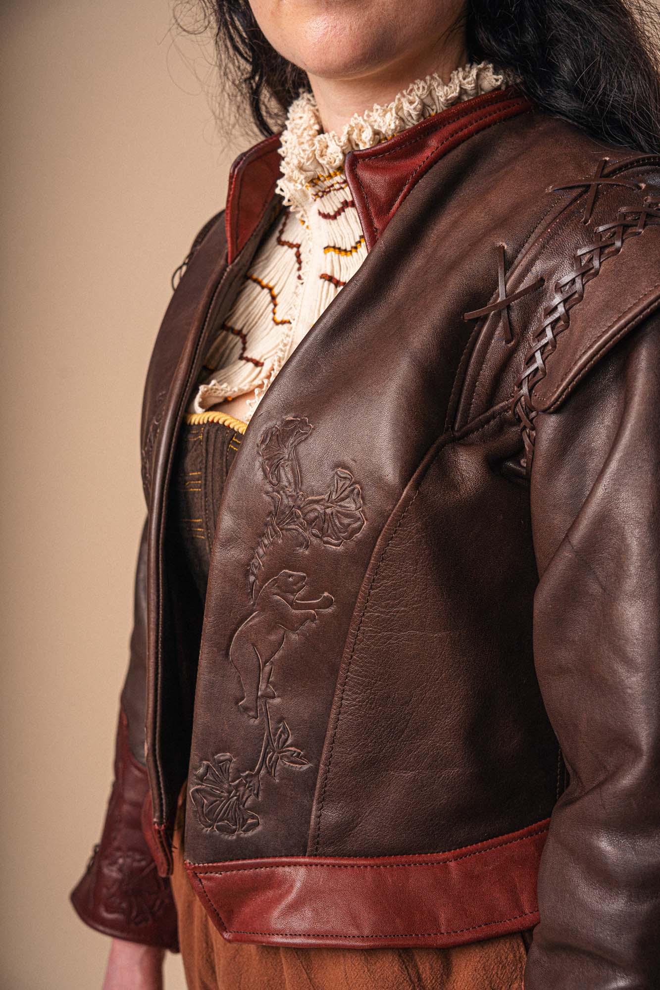 A person wearing a dark brown leather jacket with animal and floral embossing, over a cream and brown textured blouse. Only the lower half of the face is visible; background is plain beige.