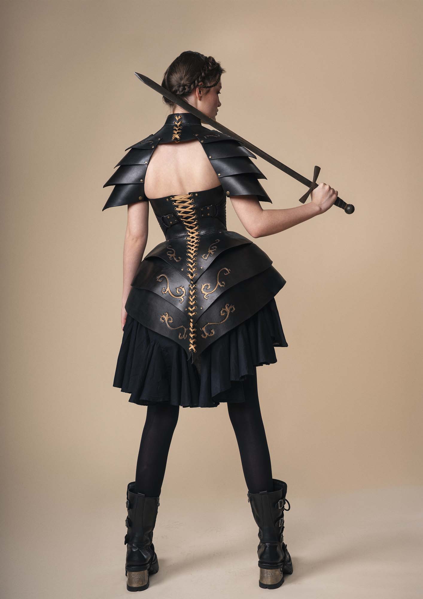 A person stands facing away, holding a sword over their shoulder. They wear black layered armour with gold accents, a black dress, black tights, and black boots, against a beige background.