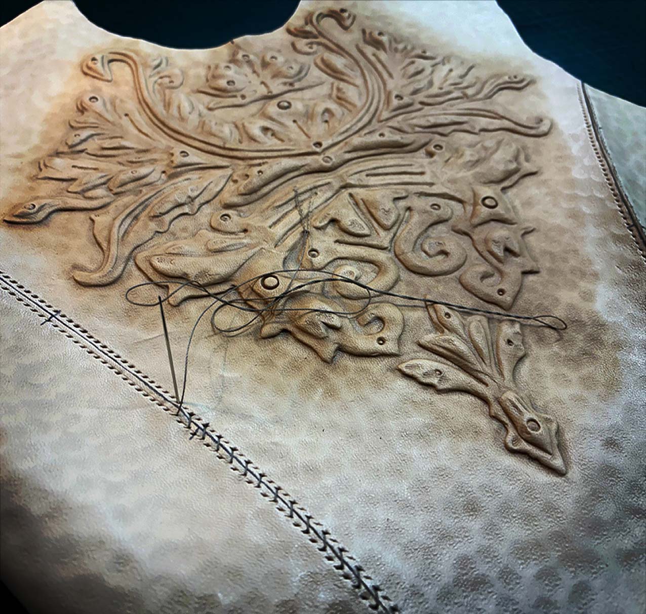 A close-up of a piece of light tan leather with intricate embossed patterns and visible dark thread, showing part of a decorative hand-stitching process along the edge.