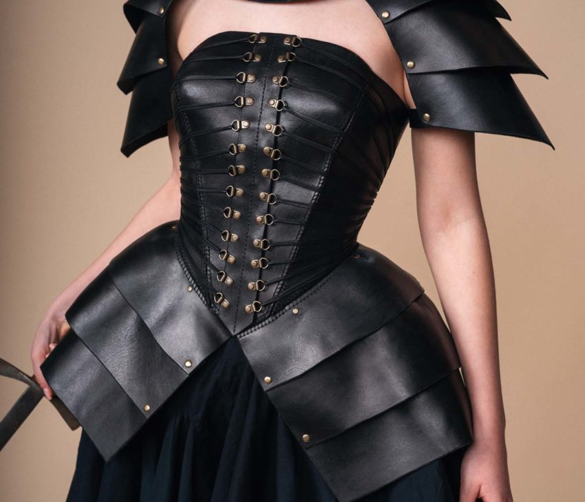 Person wearing black leather armour with layered shoulder plates, corset-style bodice with lacing and eyelets, and a skirt. Armour features a gold emblem at the neck. Background is beige. Face is partially out of frame.