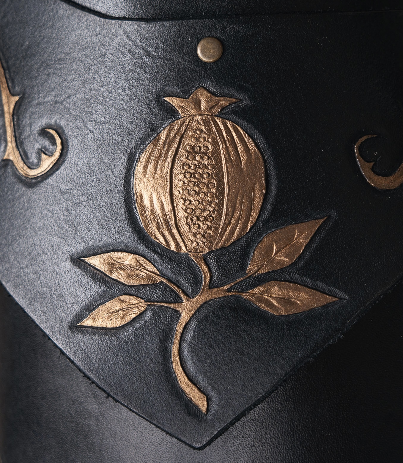 A close-up of a dark leather surface with a raised, gold-coloured pomegranate design and leaves. The detailed embossing is centred on a pointed section of the leather.