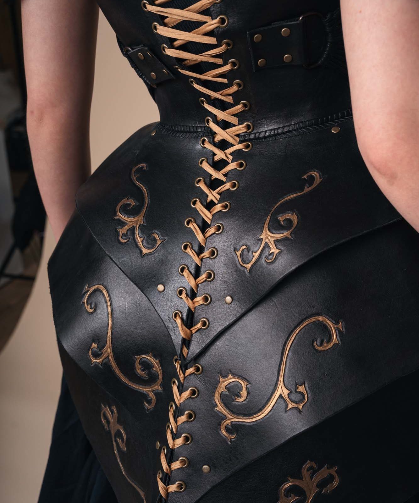 A close-up of a person wearing a black corset with tan laces and gold ornamental designs. The back of the corset features layered leather panels and decorative patterns. Only the torso and part of the upper arms are visible.