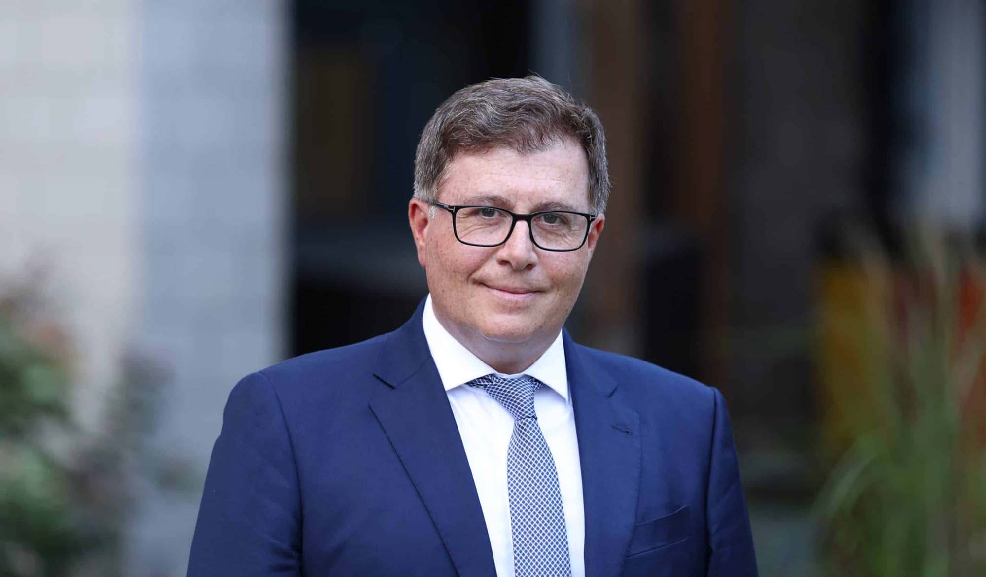 A man with short brown hair and glasses wearing a navy blue suit, white shirt, and patterned blue tie.